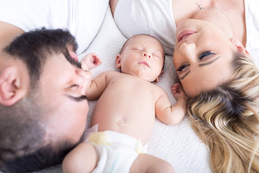 Two light skinned parents lay on either side of a sleeping newborn baby.